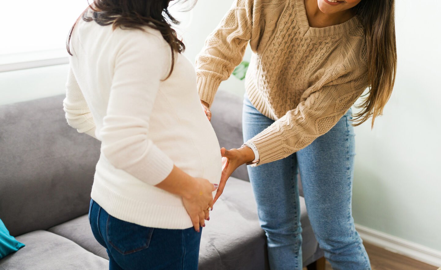 Attractive hispanic midwife smiling while touching the round belly of a caucasian pregnant woman in her home