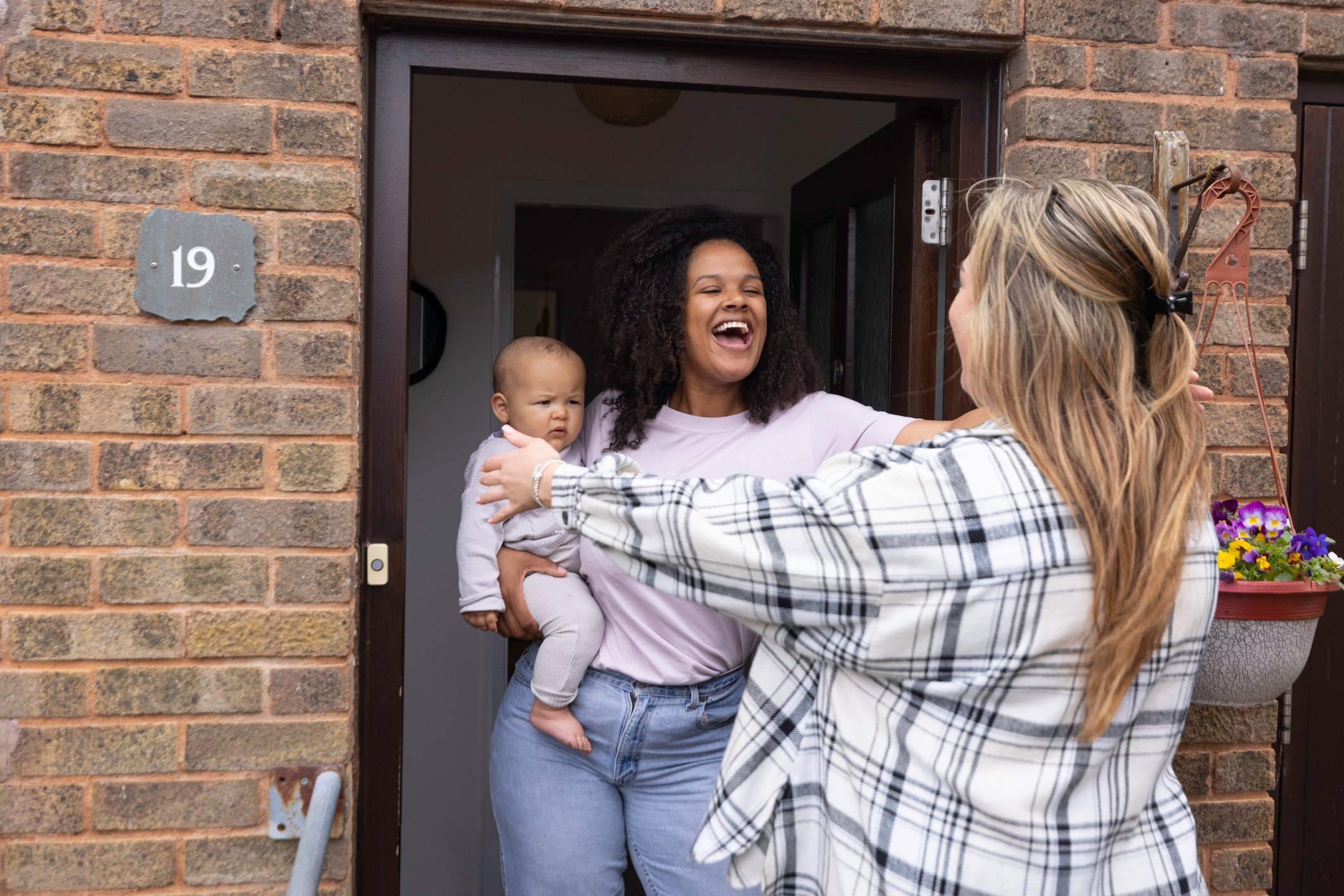 A multiracial mother holding her baby whilst standing in her doorway goes to hug and greet her friend.