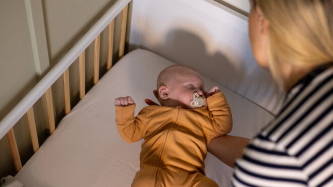 Close-up over-the-shoulder shot of a mother holding her young baby boy as she begins to place him in his cot. The woman is wearing a striped top. The baby is sleeping with a dummy in his mouth.